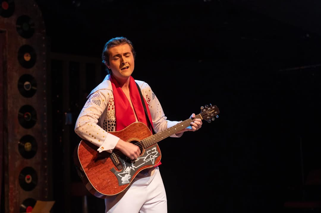 Tristan singing and playing guitar on stage in an Elvis-style costume